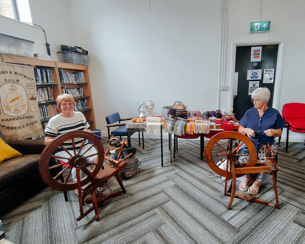 Two members with their spinning wheels, in front of a table showing woven and knitted items.