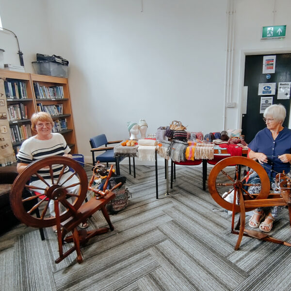 Two members with their spinning wheels, in front of a table showing woven and knitted items.