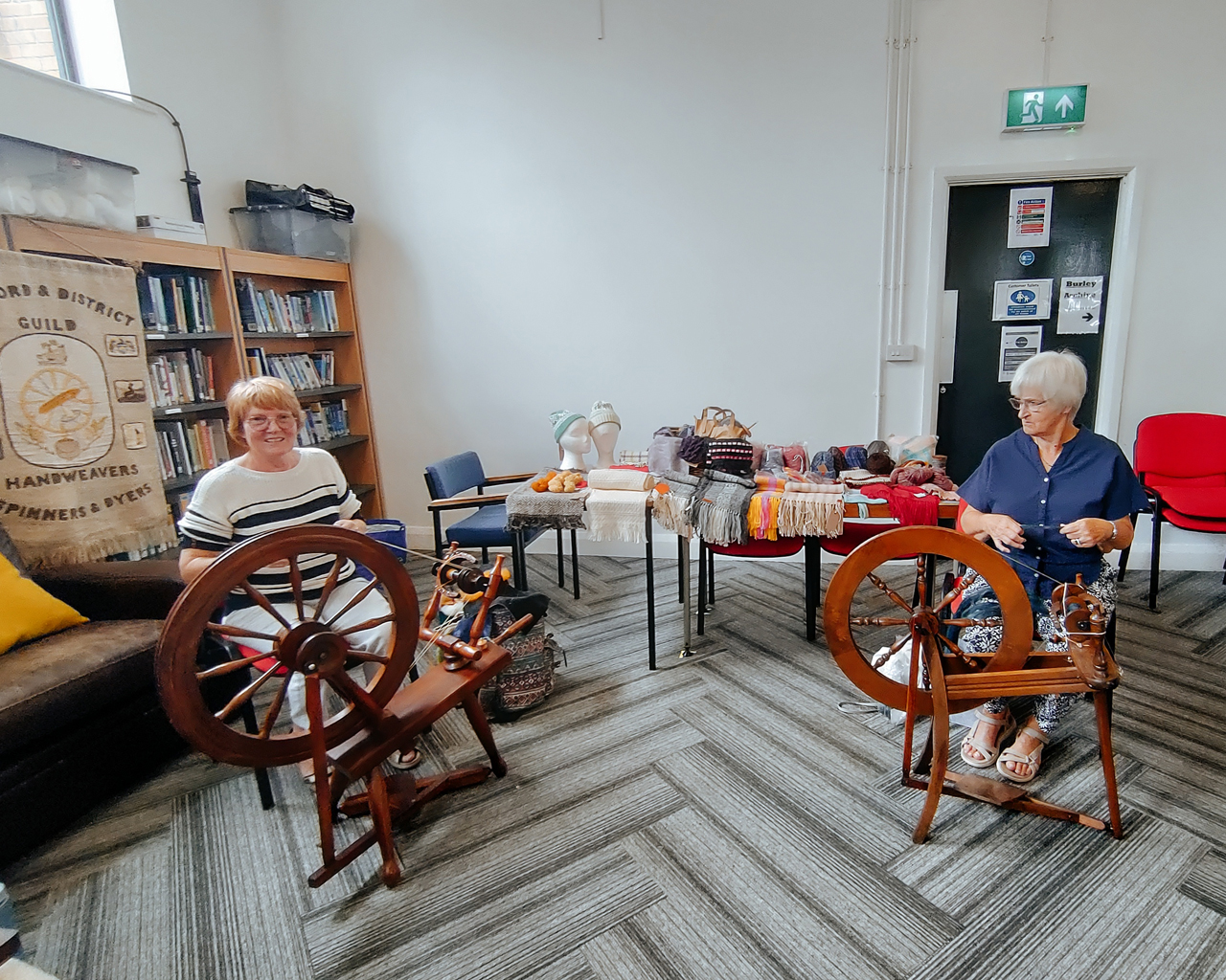 Two members with their spinning wheels, in front of a table showing woven and knitted items.