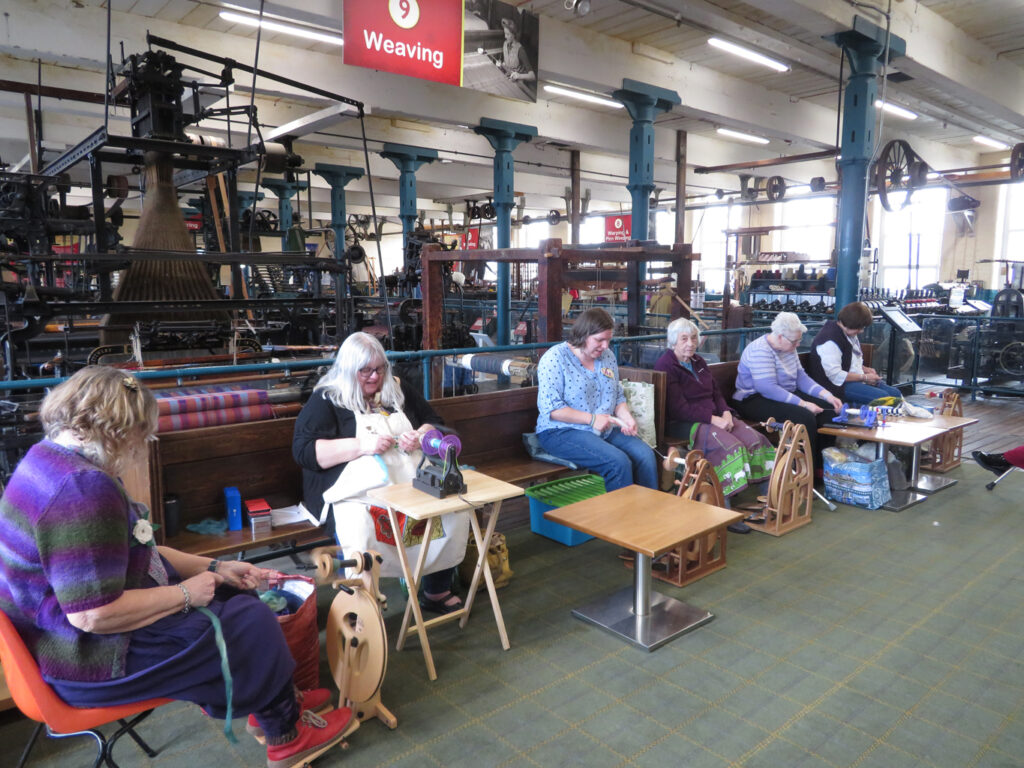 Members of BWSD with their spinning wheels, with historic looms behind them.