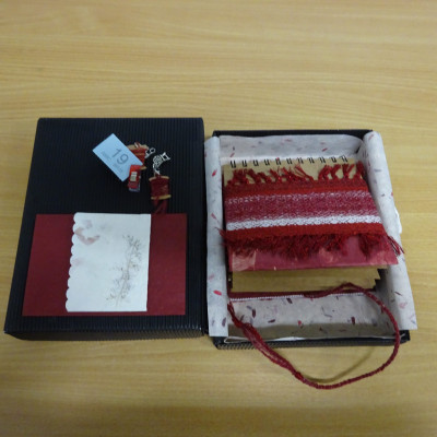Small weaving in red and white. part of a ring-bound book, in an open box.