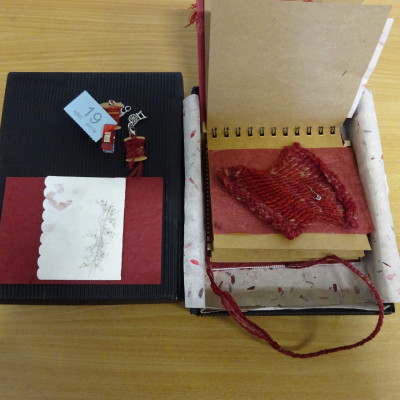 Small knitted sample, in red. Part of a ring-bound book, in an open box.