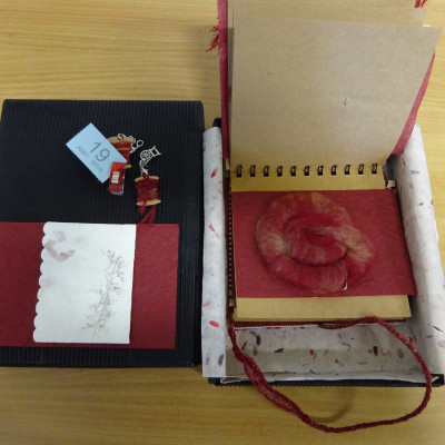 Coiled rolag of wool, in red and white. Part of a ring-bound book, in an open box.