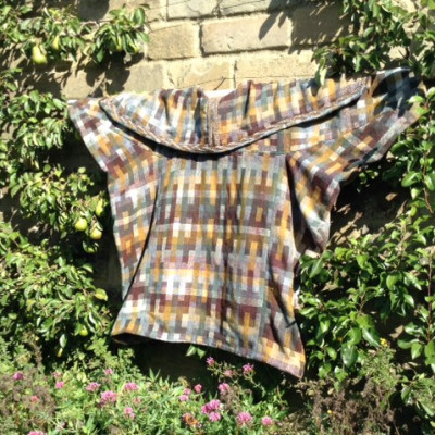 A yellow, grey and cream coat hung on a wall between two bushes.